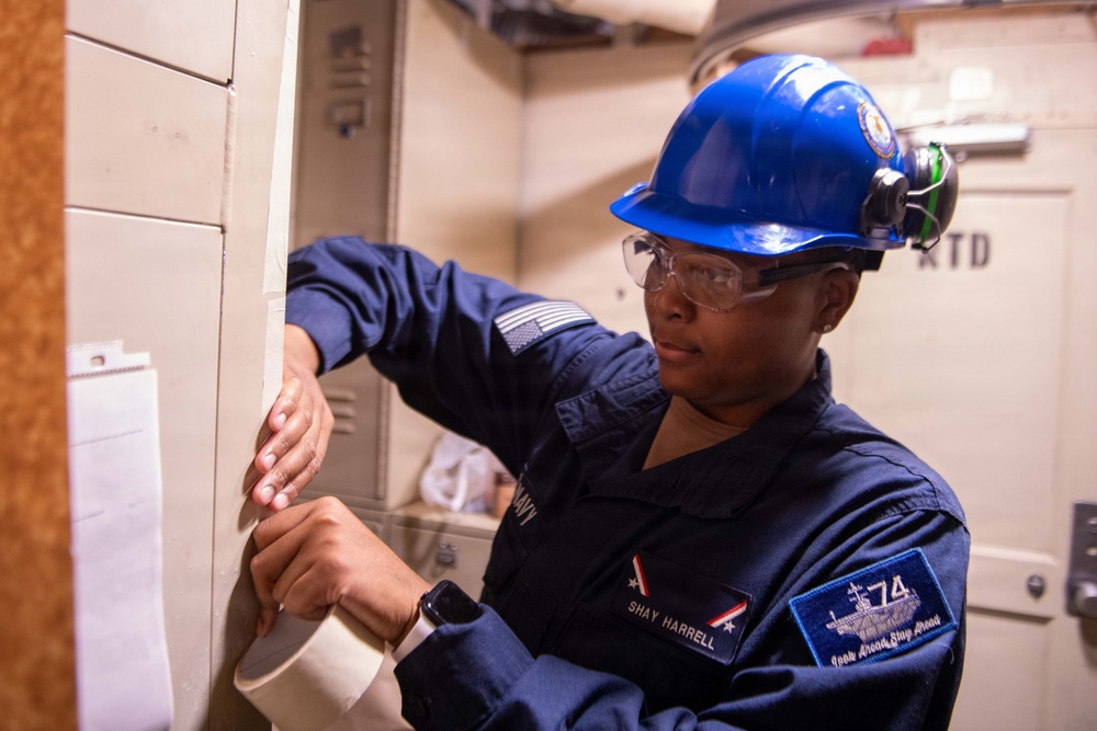 A U.S. Navy sailor named Shay Harrell is performing maintenance or inspection work in what appears to be a shipboard compartment. She is wearing standard Navy coveralls, a blue hard hat, and safety glasses while working with her hands on a panel or wall surface. The patch on her sleeve indicates she is assigned to a vessel designated '74', consistent with USS John C. Stennis (CVN-74).