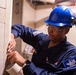 A U.S. Navy sailor named Shay Harrell is performing maintenance or inspection work in what appears to be a shipboard compartment. She is wearing standard Navy coveralls, a blue hard hat, and safety glasses while working with her hands on a panel or wall surface. The patch on her sleeve indicates she is assigned to a vessel designated '74', consistent with USS John C. Stennis (CVN-74).