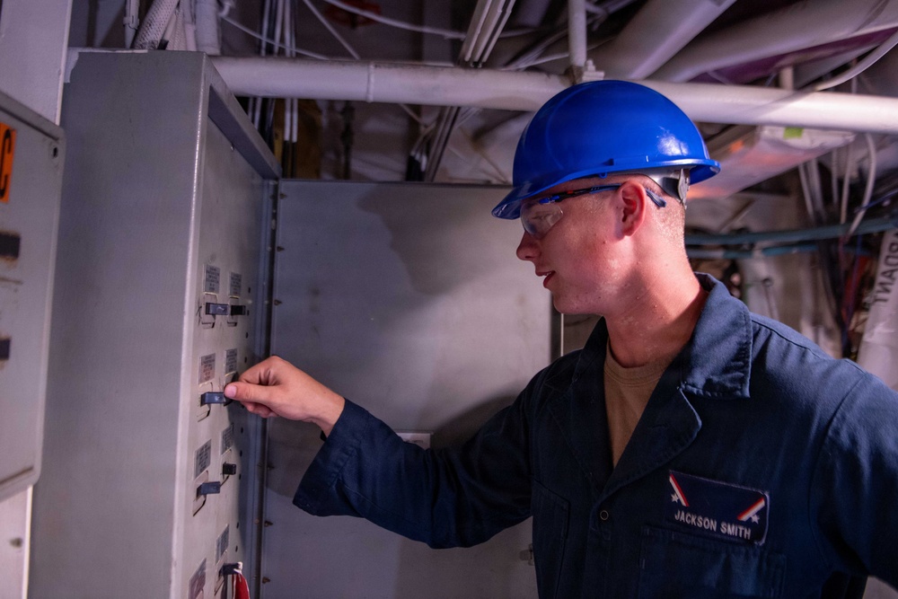 A worker identified as 'Jackson Smith' wearing a blue hard hat and safety glasses is inspecting or operating an electrical panel or control box in what appears to be an industrial or naval vessel environment. He is dressed in a dark blue uniform/coverall with a name tag visible on his chest. The setting shows overhead pipes, conduits, and cables typical of a ship's interior or industrial facility.