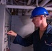 A worker identified as 'Jackson Smith' wearing a blue hard hat and safety glasses is inspecting or operating an electrical panel or control box in what appears to be an industrial or naval vessel environment. He is dressed in a dark blue uniform/coverall with a name tag visible on his chest. The setting shows overhead pipes, conduits, and cables typical of a ship's interior or industrial facility.