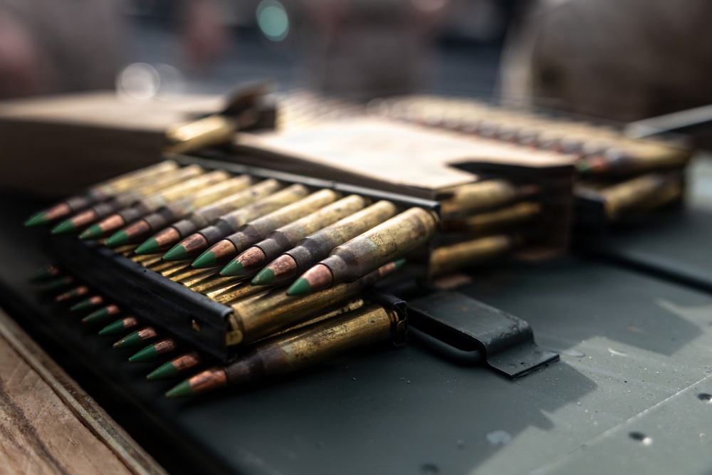 The image shows multiple rifle cartridges/rounds arranged in stripper clips on what appears to be a metal surface, likely an ammunition can or military equipment. The rounds feature distinctive green-tipped bullets, consistent with armor-piercing or penetrator ammunition types. The cartridges show signs of age or use, with oxidation visible on the brass casings.