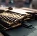 The image shows multiple rifle cartridges/rounds arranged in stripper clips on what appears to be a metal surface, likely an ammunition can or military equipment. The rounds feature distinctive green-tipped bullets, consistent with armor-piercing or penetrator ammunition types. The cartridges show signs of age or use, with oxidation visible on the brass casings.