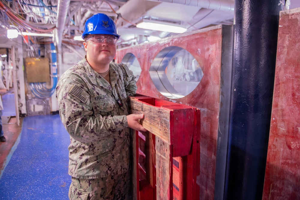 A U.S. Navy sailor in NWU Type III camouflage uniform and blue hard hat stands inside what appears to be a naval vessel's interior workspace. The sailor, whose name tape reads 'KRALOVEE' (or similar), is posing next to a red-painted wooden and metal structure with circular openings, likely a damage control or maintenance fixture. The setting appears to be a ship's lower deck or hangar bay area with overhead piping, wiring, and fluorescent lighting visible.