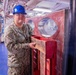 A U.S. Navy sailor in NWU Type III camouflage uniform and blue hard hat stands inside what appears to be a naval vessel's interior workspace. The sailor, whose name tape reads 'KRALOVEE' (or similar), is posing next to a red-painted wooden and metal structure with circular openings, likely a damage control or maintenance fixture. The setting appears to be a ship's lower deck or hangar bay area with overhead piping, wiring, and fluorescent lighting visible.