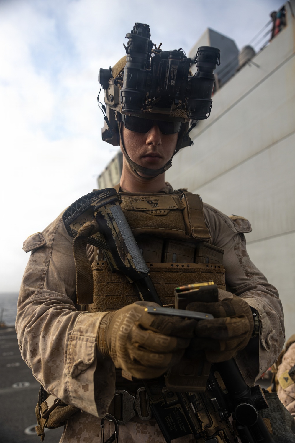 A U.S. Marine in full combat gear stands on what appears to be the deck of a naval vessel. The Marine is equipped with night vision goggles mounted on a tactical helmet and is handling what appears to be a pistol with visible ammunition rounds. The setting suggests a maritime security or visit, board, search, and seizure (VBSS) operation.