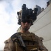 A U.S. Marine in full combat gear stands on what appears to be the deck of a naval vessel. The Marine is equipped with night vision goggles mounted on a tactical helmet and is handling what appears to be a pistol with visible ammunition rounds. The setting suggests a maritime security or visit, board, search, and seizure (VBSS) operation.