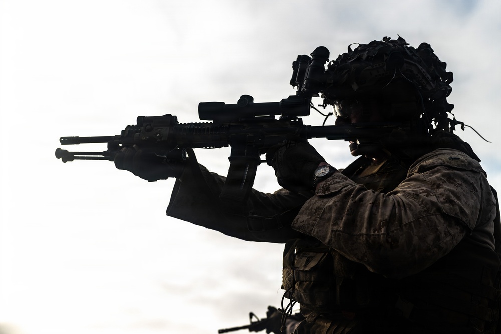 A silhouetted military operator is photographed in profile against an overcast sky, aiming a rifle equipped with optical and tactical accessories. The figure is wearing full combat gear including a tactical helmet with mounted devices, body armor, and camouflage uniform. The dramatic backlighting creates a high-contrast silhouette effect.