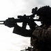 A silhouetted military operator is photographed in profile against an overcast sky, aiming a rifle equipped with optical and tactical accessories. The figure is wearing full combat gear including a tactical helmet with mounted devices, body armor, and camouflage uniform. The dramatic backlighting creates a high-contrast silhouette effect.