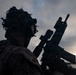 A silhouetted military personnel is photographed against an overcast sky at dusk or dawn. The individual is equipped with advanced tactical gear and is holding an assault rifle in a ready position. The dramatic backlighting creates a high-contrast silhouette effect.