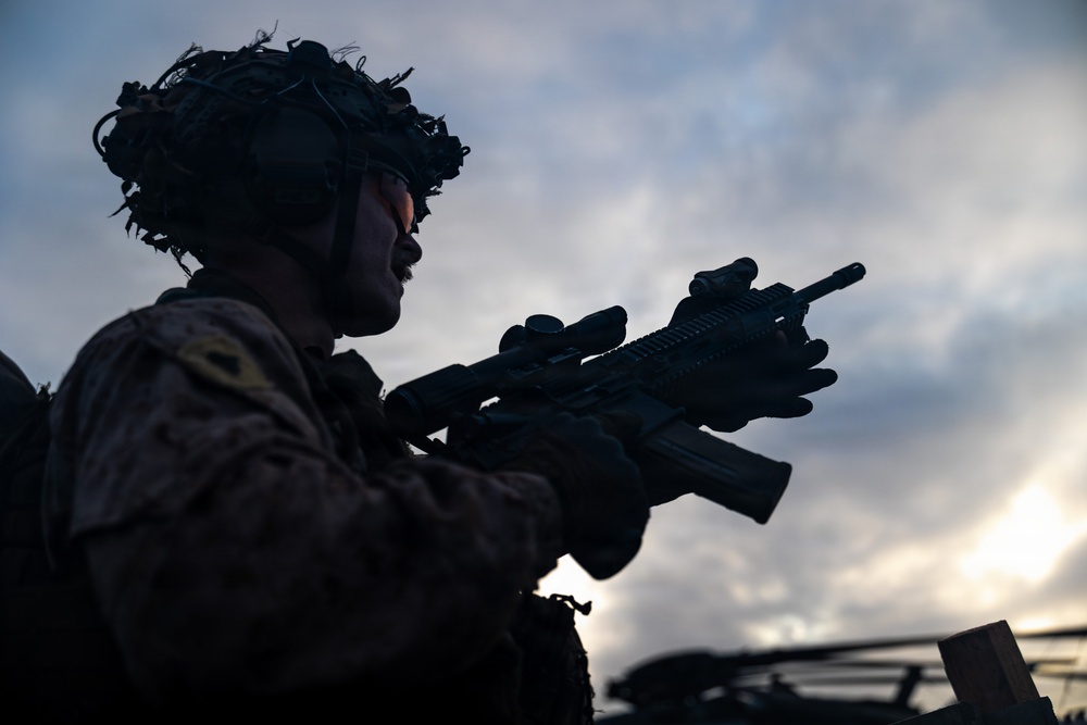 A silhouetted military personnel is photographed against a dramatic overcast sky at dusk or dawn. The soldier is equipped with full combat gear including a helmet with mounted devices and is holding an assault rifle with optical attachments. The image is taken from a low angle, emphasizing the dramatic lighting and tactical equipment.