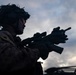 A silhouetted military personnel is photographed against a dramatic overcast sky at dusk or dawn. The soldier is equipped with full combat gear including a helmet with mounted devices and is holding an assault rifle with optical attachments. The image is taken from a low angle, emphasizing the dramatic lighting and tactical equipment.