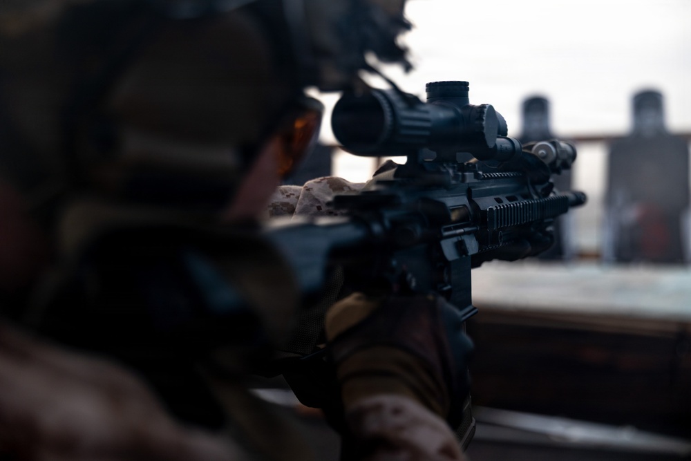 A military or law enforcement personnel is shown in a prone or low shooting position, aiming a rifle equipped with an optical scope downrange. The shooter is wearing camouflage uniform and a tactical helmet, suggesting a training or operational context. Target silhouettes are visible in the background, indicating a shooting range environment.