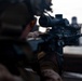 A military or law enforcement personnel is shown in a prone or low shooting position, aiming a rifle equipped with an optical scope downrange. The shooter is wearing camouflage uniform and a tactical helmet, suggesting a training or operational context. Target silhouettes are visible in the background, indicating a shooting range environment.