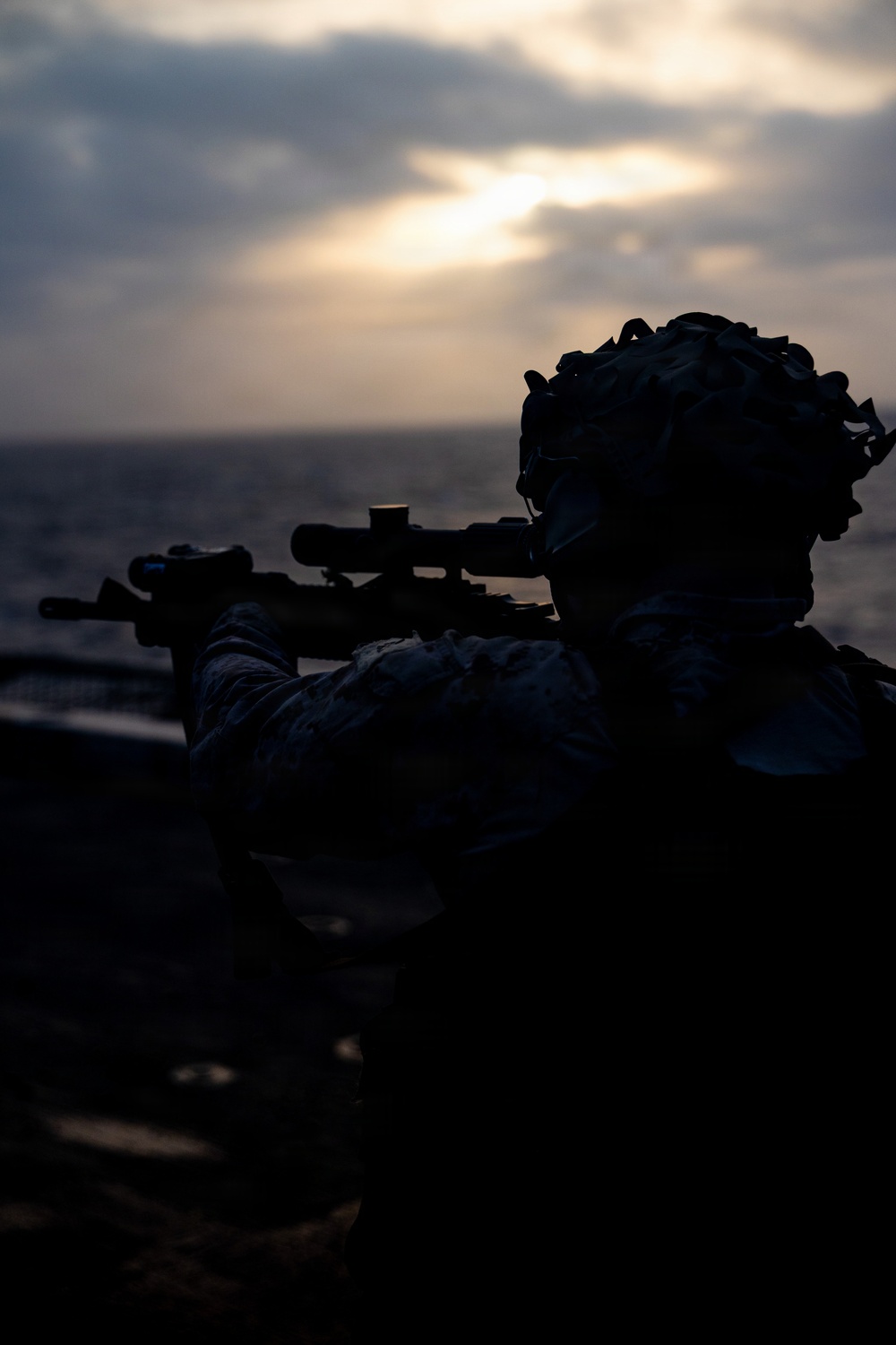 A silhouetted military personnel is shown in a prone or low-profile position aiming a scoped rifle toward the horizon over open water. The image is taken at dusk or dawn with diffused sunlight breaking through overcast clouds. The dramatic backlighting creates a strong silhouette effect, obscuring fine details of the subject.