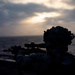 A silhouetted military personnel is shown in a prone or low-profile position aiming a scoped rifle toward the horizon over open water. The image is taken at dusk or dawn with diffused sunlight breaking through overcast clouds. The dramatic backlighting creates a strong silhouette effect, obscuring fine details of the subject.