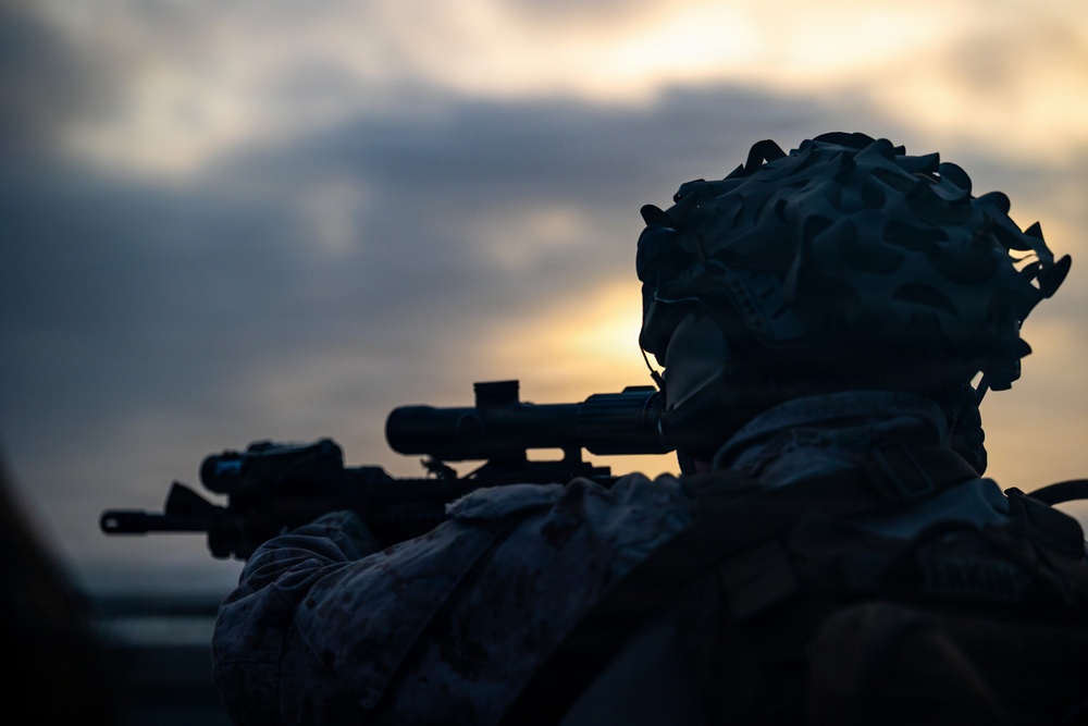 A silhouetted military personnel is shown in a prone or low-profile position aiming a scoped rifle at dusk or dawn. The figure is wearing full combat gear including a camouflage helmet with attachments, likely night vision device mounts. The dramatic backlighting from the horizon creates a high-contrast silhouette effect against a cloudy sky.