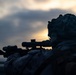 A silhouetted military personnel is shown in a prone or low-profile position aiming a scoped rifle at dusk or dawn. The figure is wearing full combat gear including a camouflage helmet with attachments, likely night vision device mounts. The dramatic backlighting from the horizon creates a high-contrast silhouette effect against a cloudy sky.
