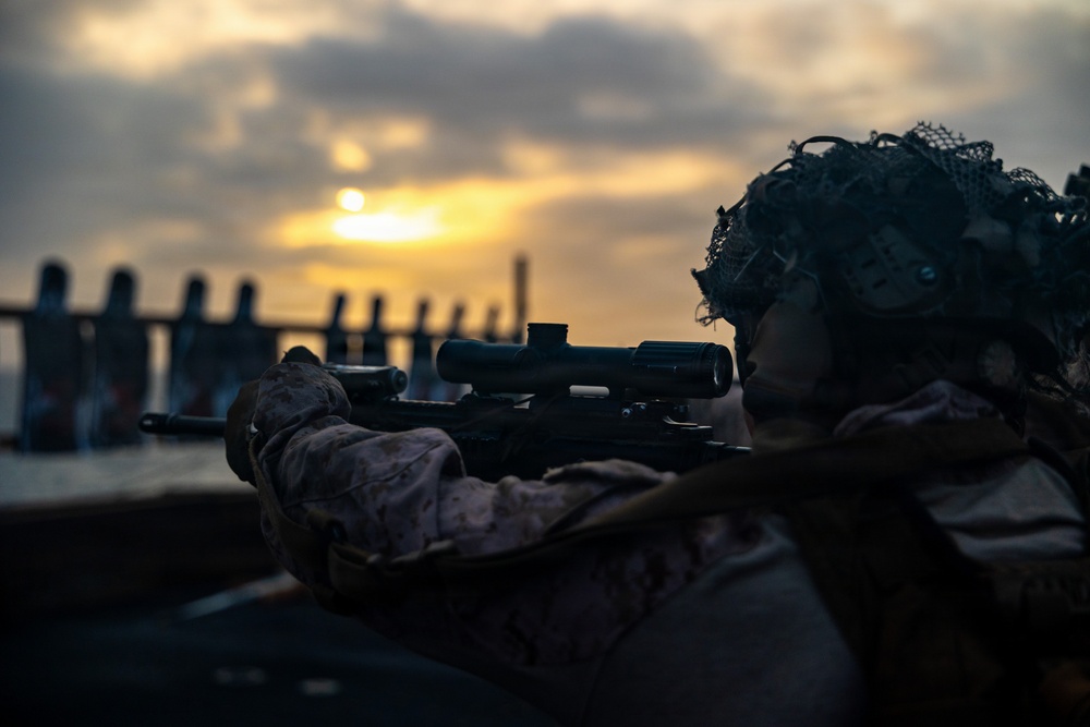 A military service member in full combat gear is shown in a prone or low firing position, aiming a scoped rifle toward a waterfront area at either dawn or dusk. The soldier is wearing MARPAT (Marine pattern) camouflage uniform and a camouflage-netted helmet, suggesting U.S. Marine Corps affiliation. The dramatic backlighting from the low sun creates a silhouette-heavy, high-contrast composition.