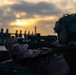 A military service member in full combat gear is shown in a prone or low firing position, aiming a scoped rifle toward a waterfront area at either dawn or dusk. The soldier is wearing MARPAT (Marine pattern) camouflage uniform and a camouflage-netted helmet, suggesting U.S. Marine Corps affiliation. The dramatic backlighting from the low sun creates a silhouette-heavy, high-contrast composition.