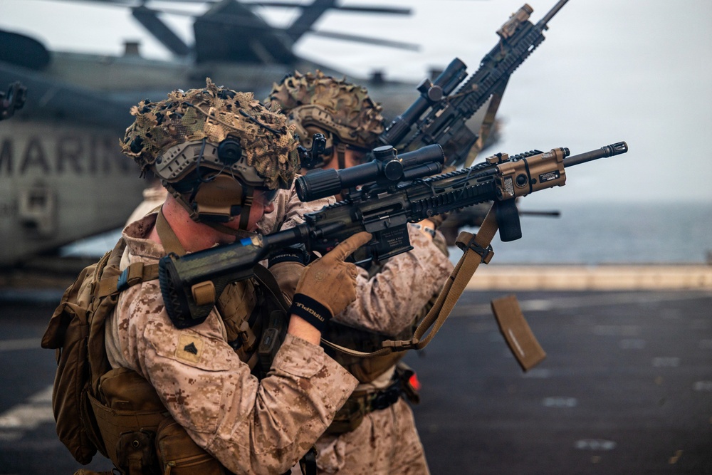 Two armed military personnel in desert camouflage uniforms are positioned on what appears to be a naval vessel's flight deck, aiming rifles in a tactical stance. The word 'MARINES' is partially visible on an aircraft in the background, identifying them as U.S. Marines. The scene appears to depict a training exercise or readiness drill aboard a ship at sea.