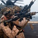 Two armed military personnel in desert camouflage uniforms are positioned on what appears to be a naval vessel's flight deck, aiming rifles in a tactical stance. The word 'MARINES' is partially visible on an aircraft in the background, identifying them as U.S. Marines. The scene appears to depict a training exercise or readiness drill aboard a ship at sea.