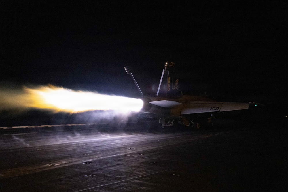 A U.S. Navy fighter jet, marked with the number '100' and tail code 'AG' with a skull-and-crossbones insignia, is captured during a night catapult launch from an aircraft carrier flight deck. The aircraft's engines are in full afterburner, producing a dramatic plume of bright white and orange flame and steam. The image was taken in near-total darkness, with the engine exhaust serving as the primary light source.