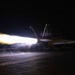 A U.S. Navy fighter jet, marked with the number '100' and tail code 'AG' with a skull-and-crossbones insignia, is captured during a night catapult launch from an aircraft carrier flight deck. The aircraft's engines are in full afterburner, producing a dramatic plume of bright white and orange flame and steam. The image was taken in near-total darkness, with the engine exhaust serving as the primary light source.