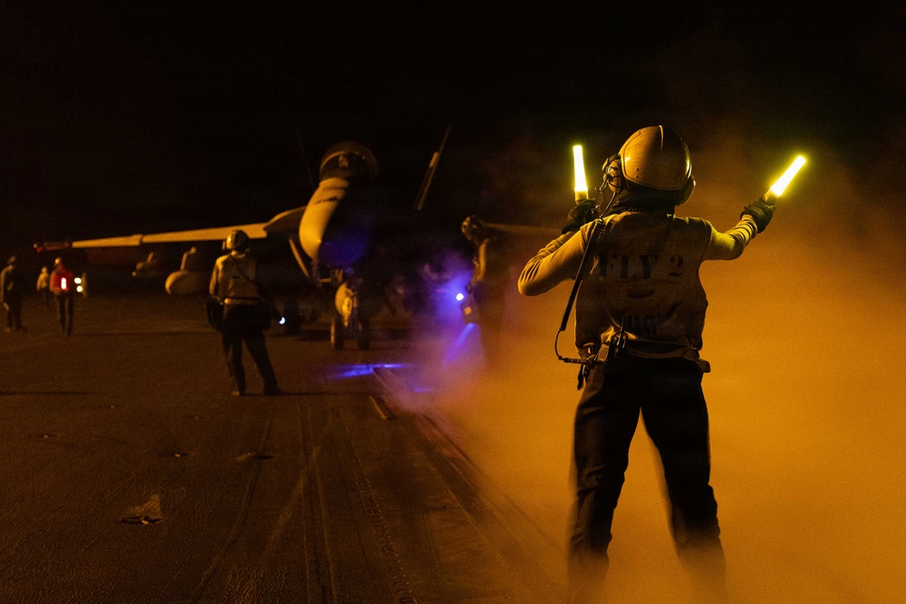 A night flight deck operation aboard what appears to be a U.S. Navy aircraft carrier, with a plane director (handler) using illuminated wands to marshal a jet aircraft. The crew member's vest is marked 'FLY 2', indicating a flight deck handling role. The scene is dramatically lit by orange haze, exhaust smoke, and blue deck lighting.
