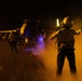 A night flight deck operation aboard what appears to be a U.S. Navy aircraft carrier, with a plane director (handler) using illuminated wands to marshal a jet aircraft. The crew member's vest is marked 'FLY 2', indicating a flight deck handling role. The scene is dramatically lit by orange haze, exhaust smoke, and blue deck lighting.