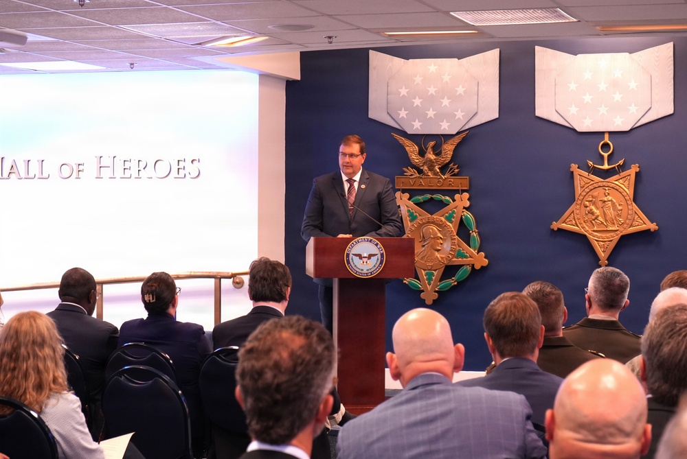 A man in a dark suit is speaking at a podium bearing the Department of Defense seal, in what appears to be the Pentagon's Hall of Heroes. The backdrop features large decorative representations of the Medal of Honor — specifically the Army and Navy versions — mounted on a dark blue wall. An audience of approximately 20 or more attendees, many in business attire and some in military uniform, are seated facing the speaker.