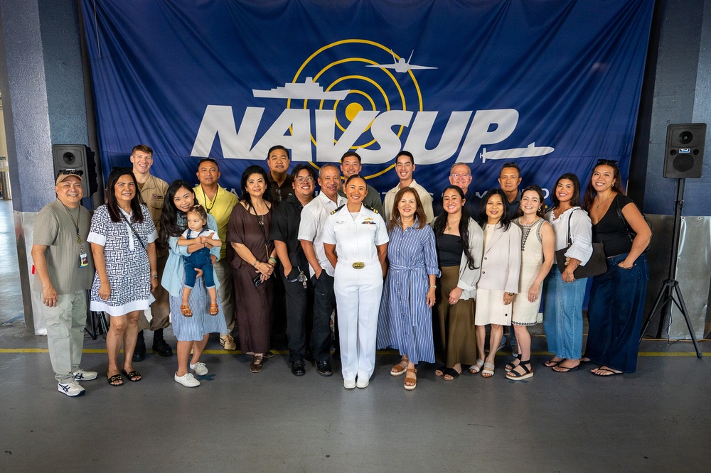 A group of approximately 20 people pose for a formal group photo in front of a large NAVSUP (Naval Supply Systems Command) banner. One individual in the center is wearing a white U.S. Navy officer's uniform, suggesting this may be a promotion, retirement, or recognition ceremony. The group appears to consist of family members, friends, and colleagues gathered to celebrate the occasion.