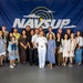 A group of approximately 20 people pose for a formal group photo in front of a large NAVSUP (Naval Supply Systems Command) banner. One individual in the center is wearing a white U.S. Navy officer's uniform, suggesting this may be a promotion, retirement, or recognition ceremony. The group appears to consist of family members, friends, and colleagues gathered to celebrate the occasion.