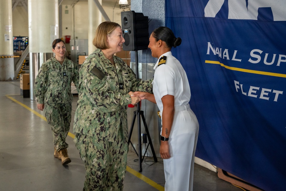 Two U.S. Navy personnel are shaking hands in what appears to be a formal military ceremony or greeting at a Naval Supply Fleet facility. One individual is wearing the Navy Working Uniform (NWU Type III camouflage) while the other is in Service Dress White uniform, indicating different duty contexts or ranks. A third service member in camouflage uniform is visible in the background approaching the scene.