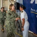 Two U.S. Navy personnel are shaking hands in what appears to be a formal military ceremony or greeting at a Naval Supply Fleet facility. One individual is wearing the Navy Working Uniform (NWU Type III camouflage) while the other is in Service Dress White uniform, indicating different duty contexts or ranks. A third service member in camouflage uniform is visible in the background approaching the scene.