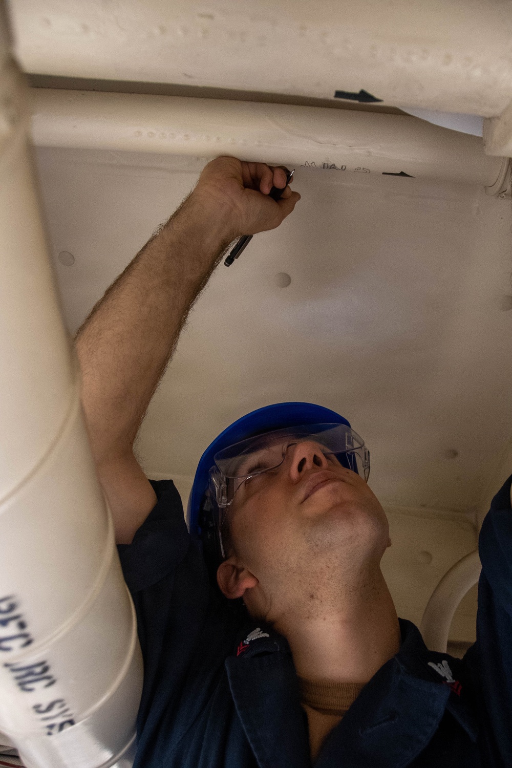 A young male worker wearing a blue hard hat and clear safety glasses is writing or marking on the underside of a white surface, likely a piece of equipment or structure. He is looking upward while holding a black marker or pen in his raised right hand. The setting appears to be an industrial or technical work environment.