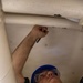 A young male worker wearing a blue hard hat and clear safety glasses is writing or marking on the underside of a white surface, likely a piece of equipment or structure. He is looking upward while holding a black marker or pen in his raised right hand. The setting appears to be an industrial or technical work environment.