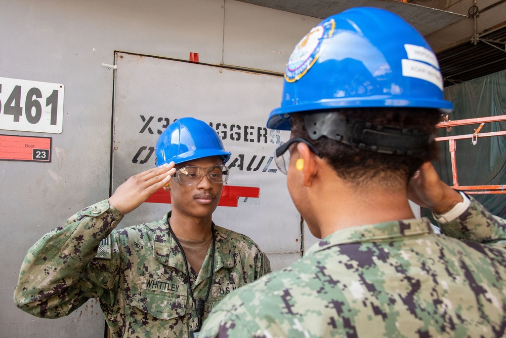 Two U.S. Navy personnel in digital camouflage uniforms and blue hard hats are exchanging salutes in what appears to be a shipyard or industrial maintenance facility. The sailor facing the camera is identified by a name tape reading 'WHITTLEY' and is wearing safety glasses. The setting suggests an active work or inspection environment with safety equipment required.