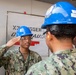 Two U.S. Navy personnel in digital camouflage uniforms and blue hard hats are exchanging salutes in what appears to be a shipyard or industrial maintenance facility. The sailor facing the camera is identified by a name tape reading 'WHITTLEY' and is wearing safety glasses. The setting suggests an active work or inspection environment with safety equipment required.