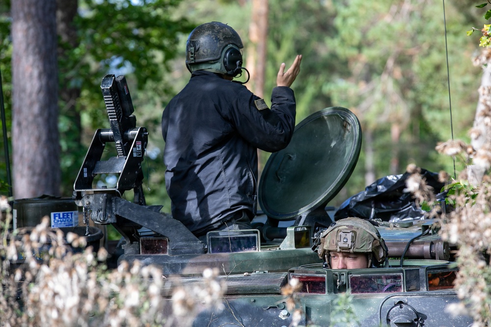 The image shows two military personnel positioned on or in what appears to be a U.S. Army armored vehicle, likely a Stryker, in a forested environment. One individual wearing a black jacket and helmet with communications headset is standing and making a hand signal, while a second soldier in standard military combat gear is visible in a lower hatch. The vehicle is partially obscured by vegetation and camouflage netting.