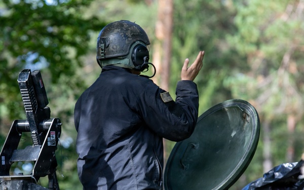 The image shows two military personnel positioned on or in what appears to be a U.S. Army armored vehicle, likely a Stryker, in a forested environment. One individual wearing a black jacket and helmet with communications headset is standing and making a hand signal, while a second soldier in standard military combat gear is visible in a lower hatch. The vehicle is partially obscured by vegetation and camouflage netting.