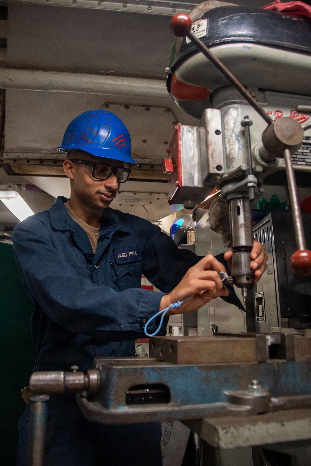 A sailor identified by name tag as 'IAGO PINA' is operating a drill press in what appears to be a shipboard machine shop. He is wearing a blue hard hat with red markings, safety glasses, and a navy blue coverall uniform. The setting shows a confined industrial workspace consistent with a naval vessel's interior.