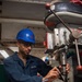 A sailor identified by name tag as 'IAGO PINA' is operating a drill press in what appears to be a shipboard machine shop. He is wearing a blue hard hat with red markings, safety glasses, and a navy blue coverall uniform. The setting shows a confined industrial workspace consistent with a naval vessel's interior.