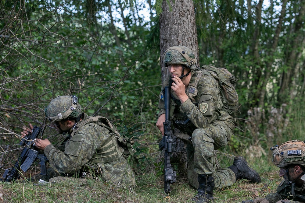 Three military personnel in camouflage uniforms are positioned in a wooded area, taking cover near trees and undergrowth. The central figure is crouching against a tree trunk while speaking into a radio handset and holding a rifle. The scene appears to depict a tactical field exercise or operational pause.
