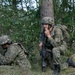 Three military personnel in camouflage uniforms are positioned in a wooded area, taking cover near trees and undergrowth. The central figure is crouching against a tree trunk while speaking into a radio handset and holding a rifle. The scene appears to depict a tactical field exercise or operational pause.