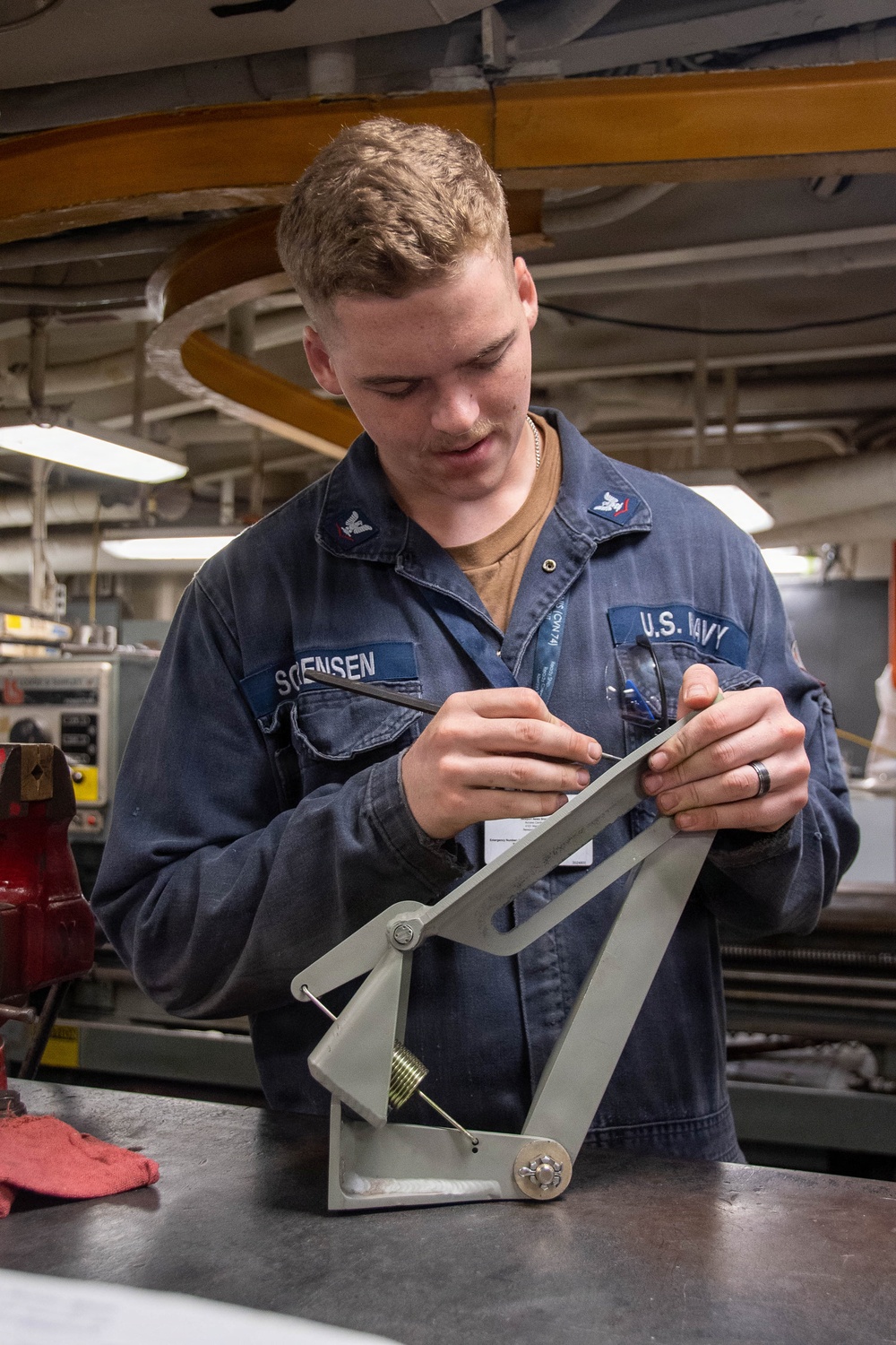 A U.S. Navy sailor identified by name tape as 'SORENSEN' is working on a mechanical component in what appears to be a shipboard machine shop or workshop. He is wearing Navy blue coveralls with 'U.S. NAVY' patch and rank insignia visible on his uniform. He is focused on assembling or adjusting a gray metal bracket/linkage assembly on a workbench.