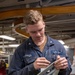 A U.S. Navy sailor identified by name tape as 'SORENSEN' is working on a mechanical component in what appears to be a shipboard machine shop or workshop. He is wearing Navy blue coveralls with 'U.S. NAVY' patch and rank insignia visible on his uniform. He is focused on assembling or adjusting a gray metal bracket/linkage assembly on a workbench.