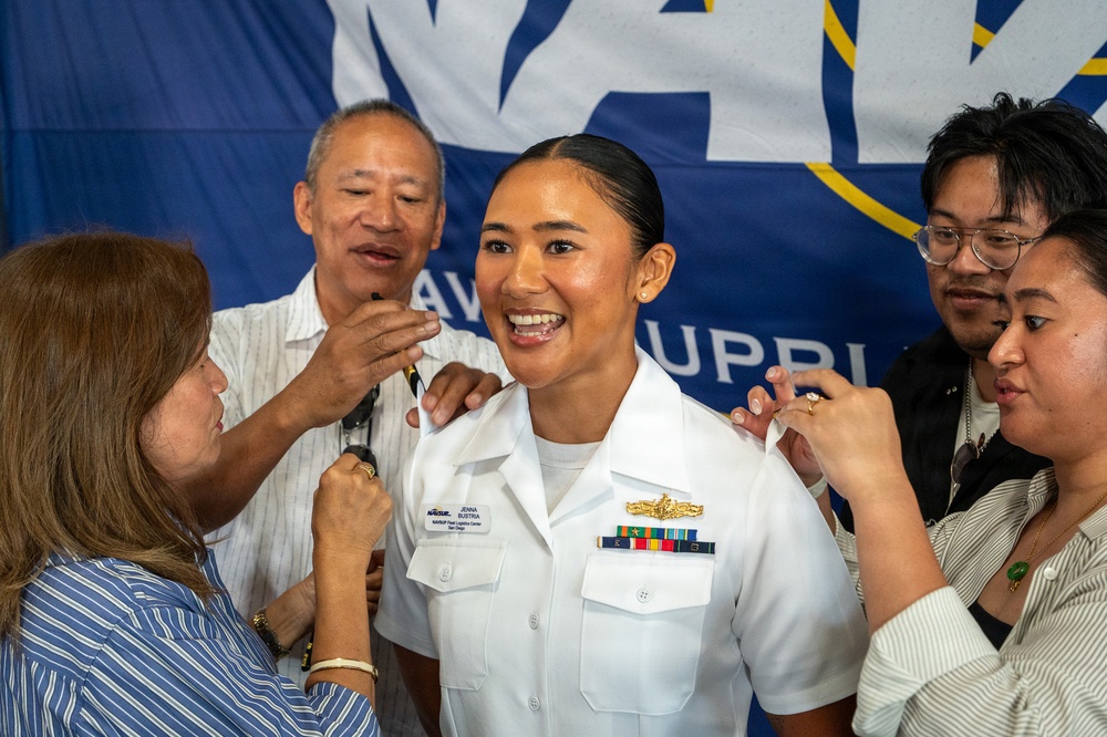 A U.S. Navy officer named Jenna Bustria is being pinned during a promotion ceremony, surrounded by what appear to be family members. She is wearing a white Navy dress uniform with ribbons and a warfare device, smiling broadly as multiple people simultaneously pin new insignia on her shoulders. A Navy banner is visible in the background.