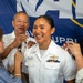 A U.S. Navy officer named Jenna Bustria is being pinned during a promotion ceremony, surrounded by what appear to be family members. She is wearing a white Navy dress uniform with ribbons and a warfare device, smiling broadly as multiple people simultaneously pin new insignia on her shoulders. A Navy banner is visible in the background.