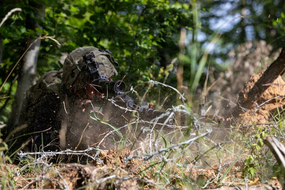 A soldier in full combat gear is in a prone firing position behind barbed wire obstacles in a wooded area. Debris, dust, and sparks are visible around the weapon, suggesting active firing. The scene appears to be a military training exercise or combat operation.