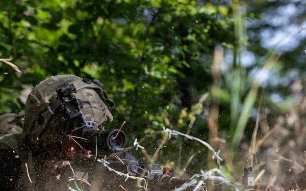A soldier in full combat gear is in a prone firing position behind barbed wire obstacles in a wooded area. Debris, dust, and sparks are visible around the weapon, suggesting active firing. The scene appears to be a military training exercise or combat operation.